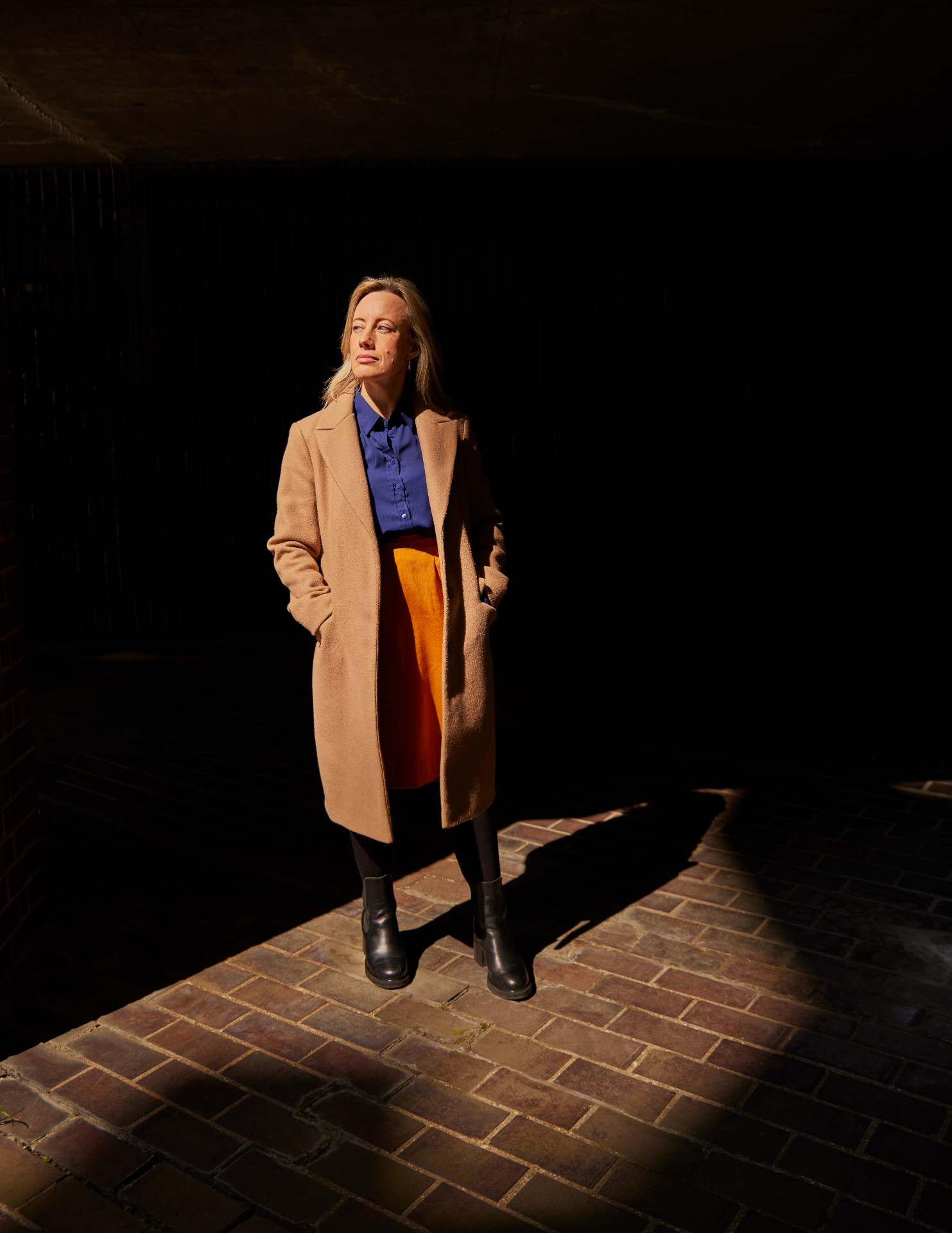Vicki Cardwell standing in a shaft of sunlight against a dark brick wall